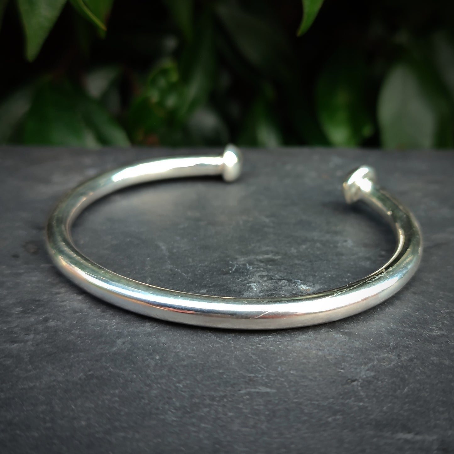 The Heavy Silver Bangle by silverhollowjewellery, crafted from recycled 925 sterling silver with rounded ends, sits on a dark stone surface with green leaves in the background.