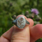 Silver ring with engraved thistle design held in a hand against a blurred natural background