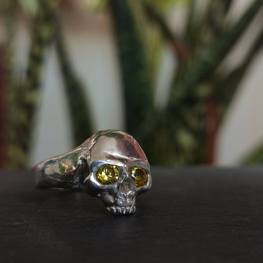 The Yellow Sapphire Skull Ring by silverhollowjewellery features yellow gemstone eyes and is crafted from recycled sterling silver, shown against a blurred green foliage background.