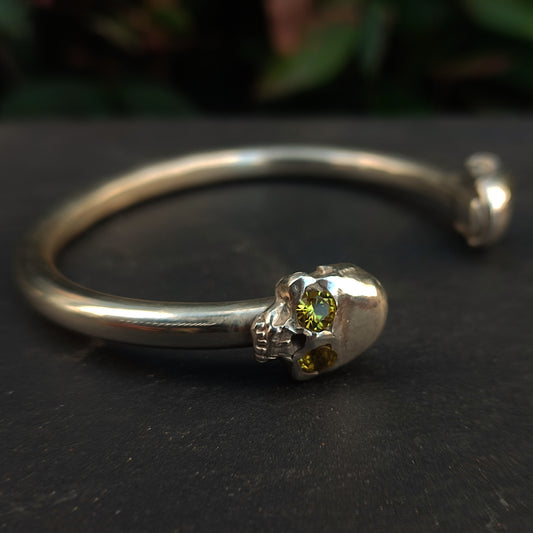 The Yellow Sapphire Skull Cuff by silverhollowjewellery is a silver cuff bracelet made from recycled sterling silver, featuring a skull design with yellow sapphires in the eyes set against a dark background.