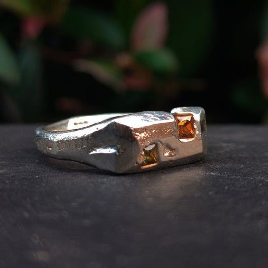The Sand Cast Sapphire Ring by silverhollowjewellery is crafted from recycled sterling silver with a textured sand-cast finish and features two square yellow-orange princess cut sapphires in an angular design.