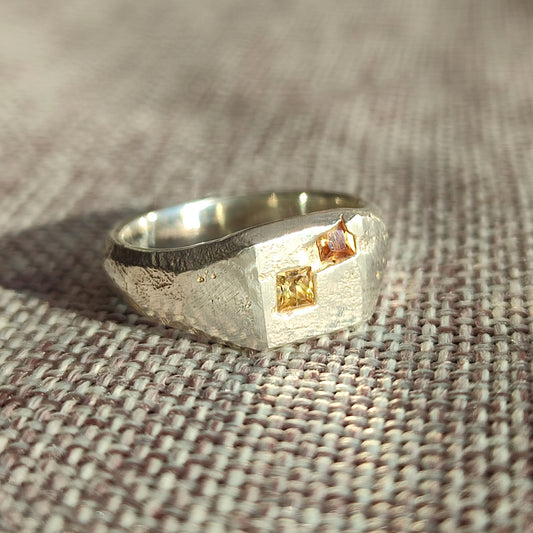 The Sand Cast Sapphire Signet by silverhollowjewellery is a recycled sterling silver ring with a textured finish, featuring two square-cut yellow and pink stones set on a flat surface.
