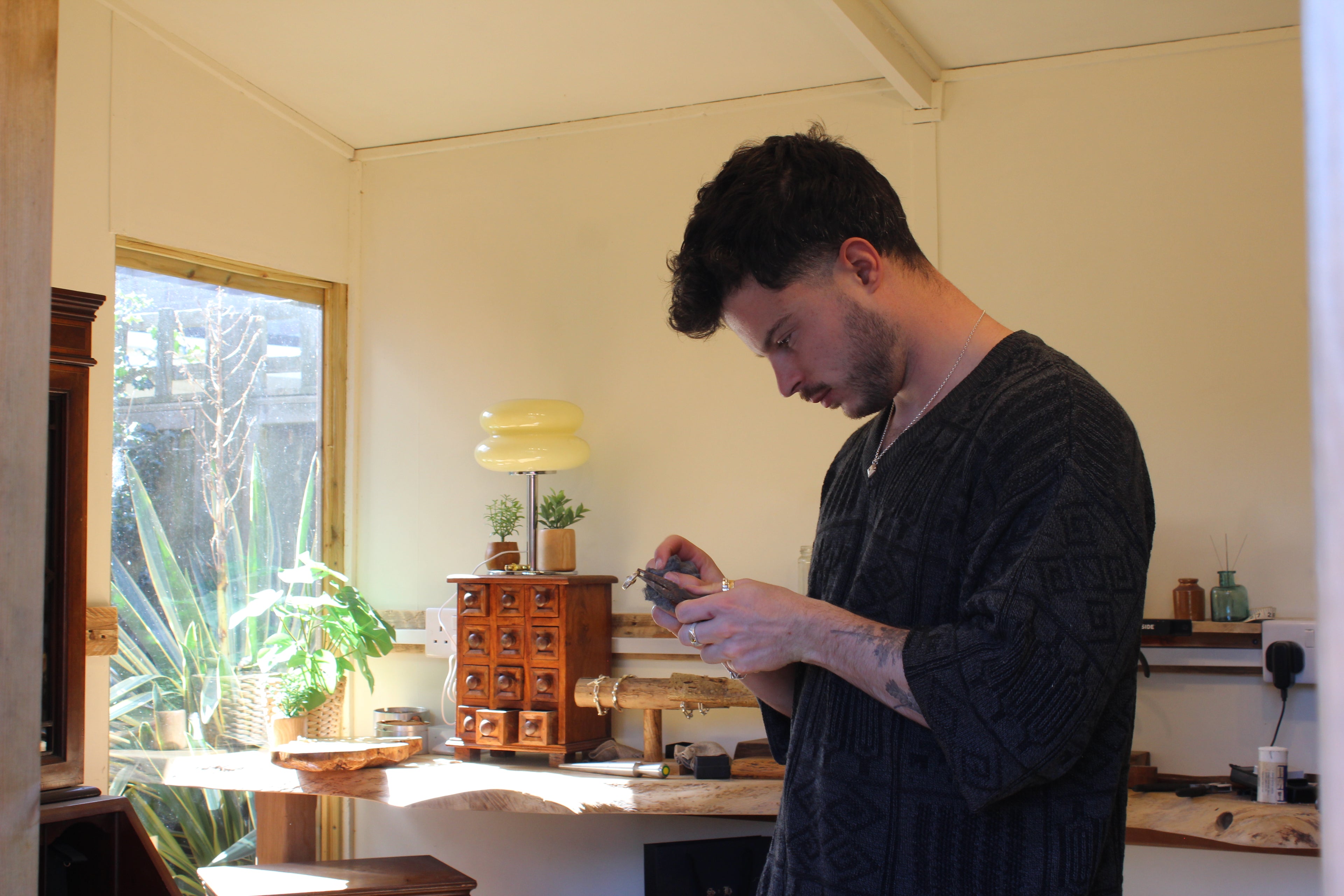 A man stands in a sunlit room examining an object in his hands, with plants and wooden shelves visible in the background.