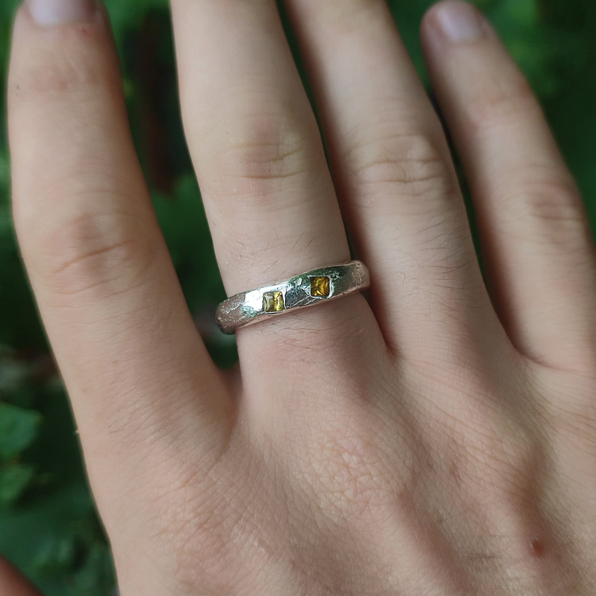 A close-up of a hand wearing a textured silver band ring with two small yellow sapphires, set against a blurred green outdoor background.