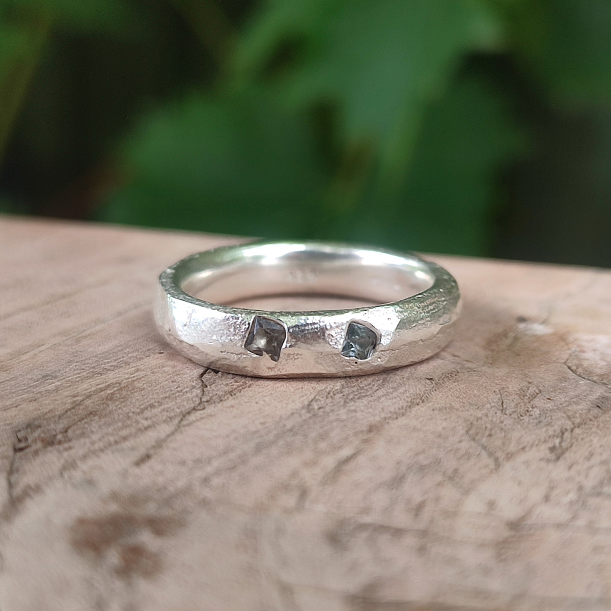 Sand cast sterling silver band ring with two flush-set teal sapphires, resting on driftwood