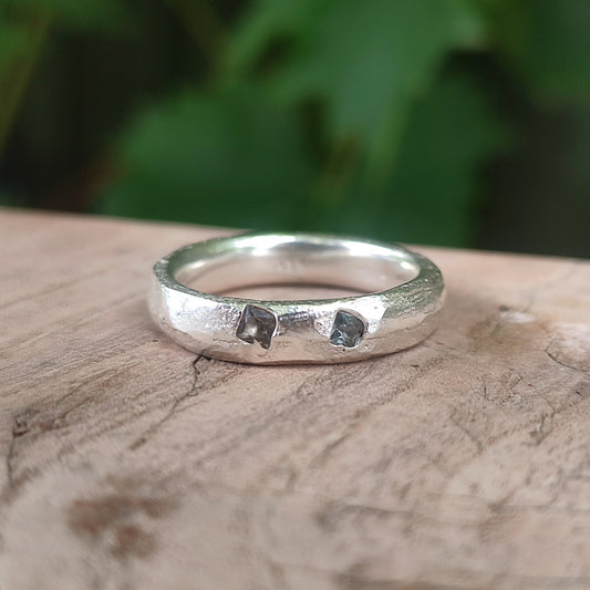 Sand cast sterling silver band ring with two flush-set teal sapphires, resting on driftwood