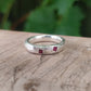 A textured sterling silver ring with two small square-cut ruby gemstones, resting on a rustic wooden surface with a blurred green background.