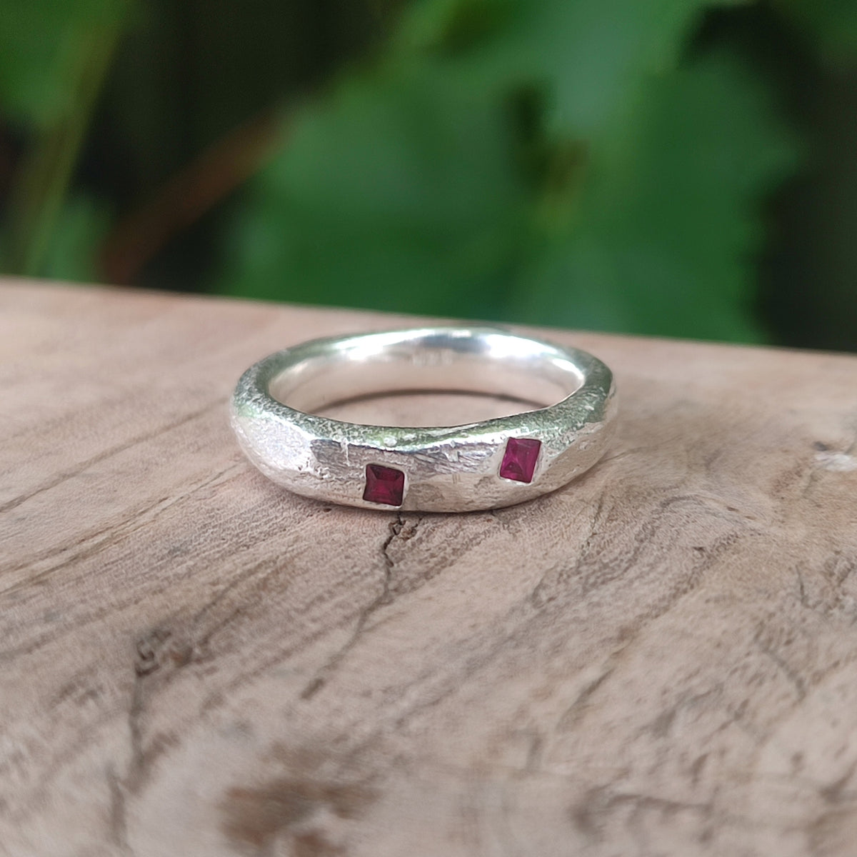 A textured sterling silver ring with two small square-cut ruby gemstones, resting on a rustic wooden surface with a blurred green background.