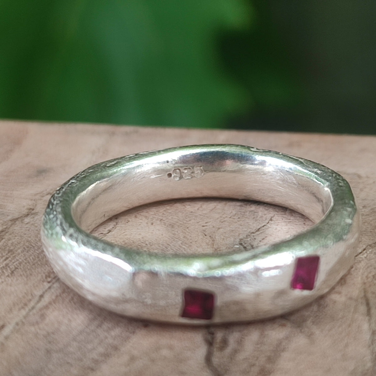 A texured sterling silver ring with two small square-cut red gemstones, with a .925 stamp on the inside, and the ring on a rustic wooden surface with a blurred green background.