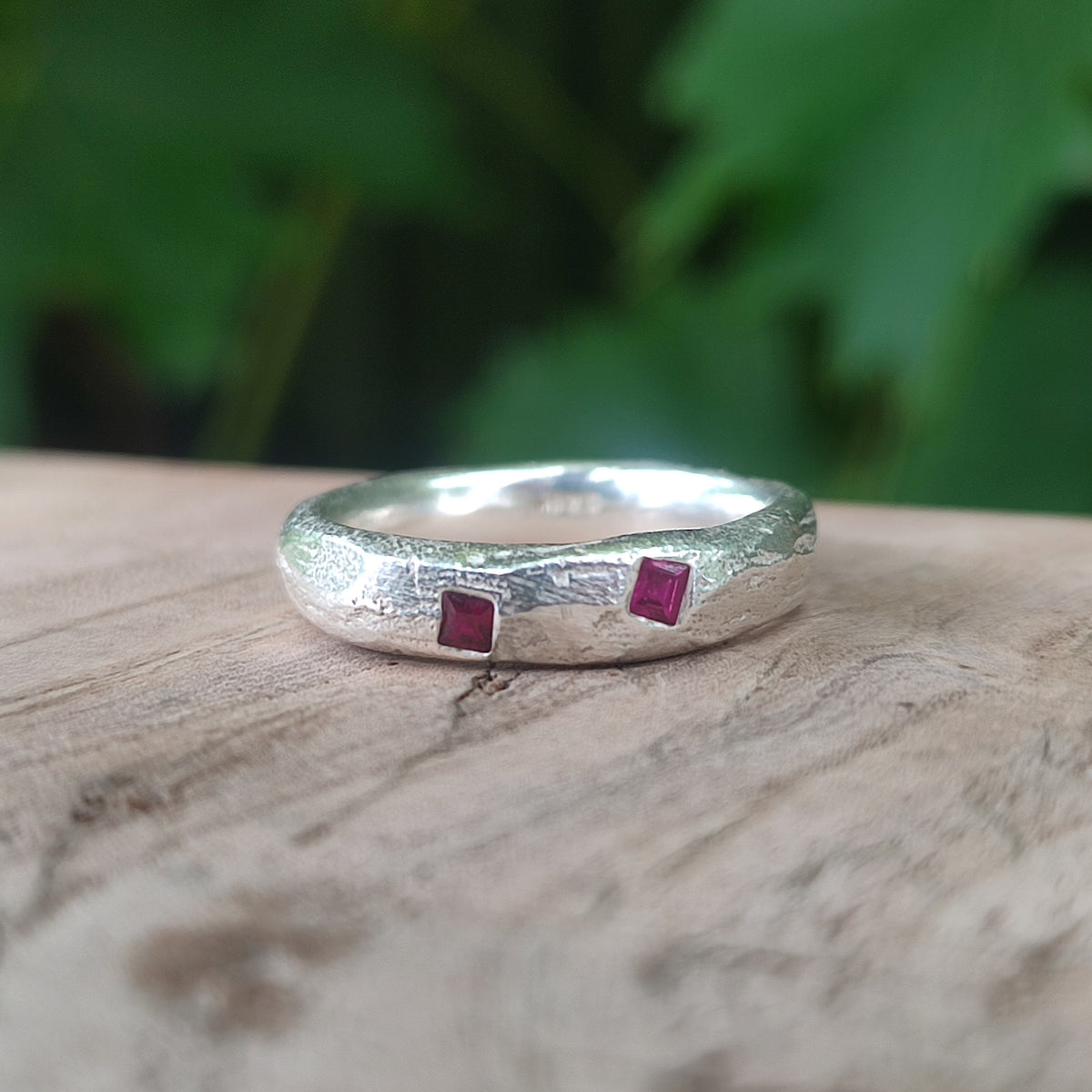 A textured sterling silver ring with two small square-cut runy gemstones, resting on a rustic wooden surface with a blurred green background.