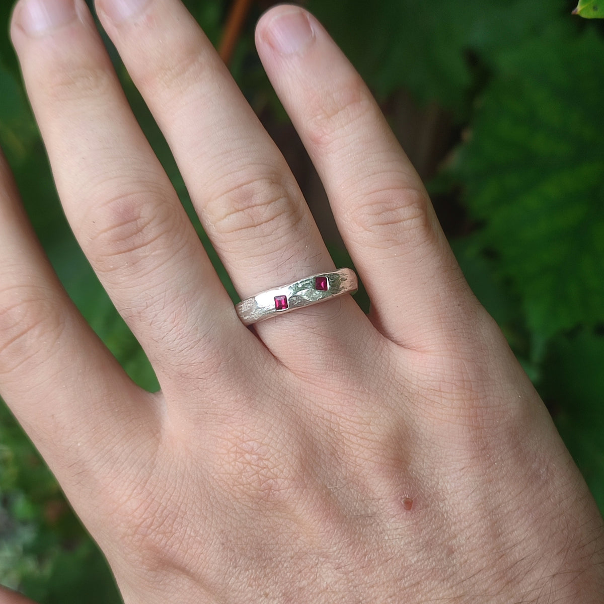 A close-up of a hand wearing a silver band ring with two small square-cut ruby gemstones, set against a blurred green outdoor background.