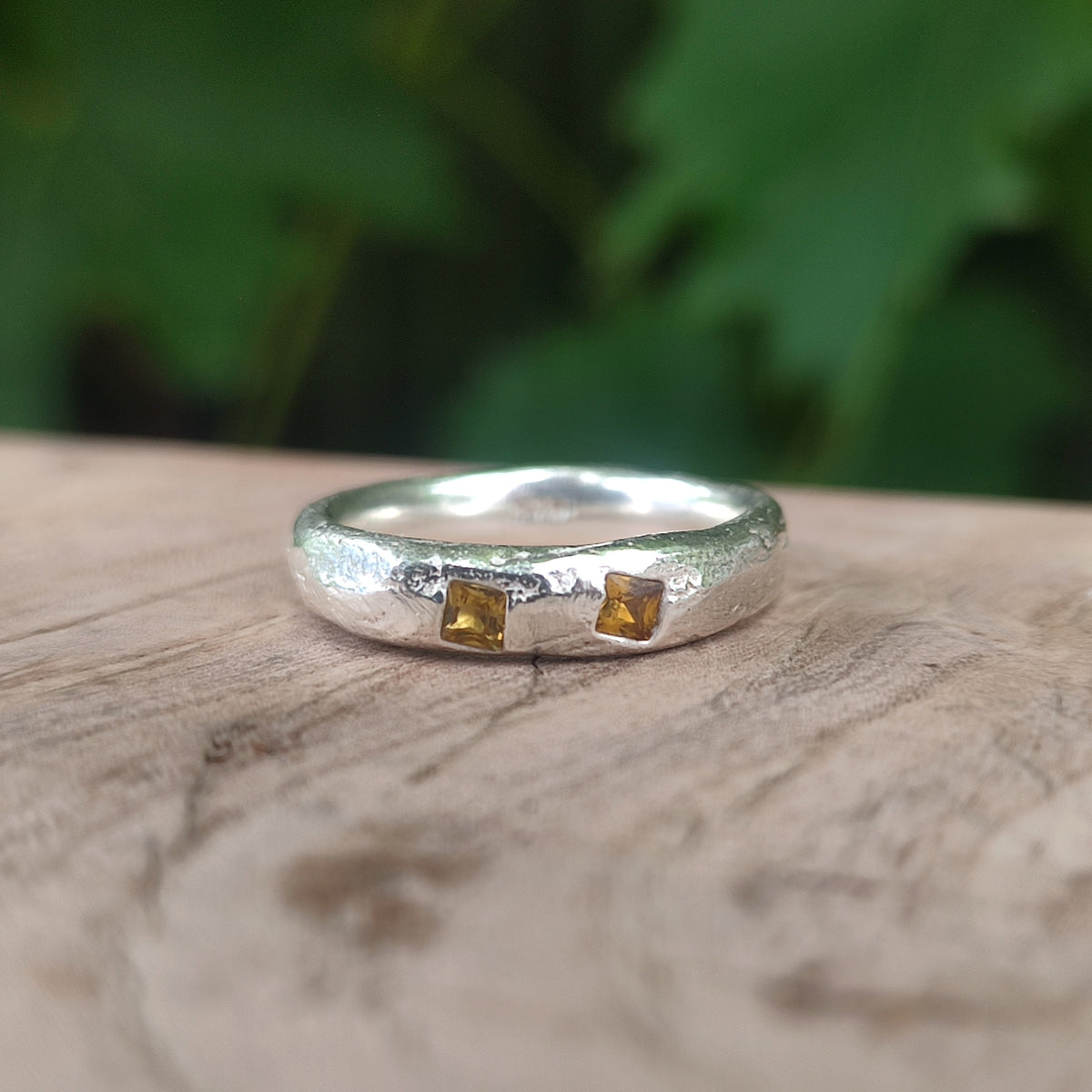 A  sterling silver ring with two square yellow sapphires, resting on a rustic wooden surface with a blurred green background.
