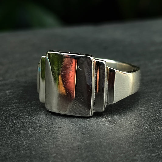 The Silver Stepped Signet Ring by silverhollowjewellery, featuring a bold geometric Art Deco design, rests on a dark, textured surface.